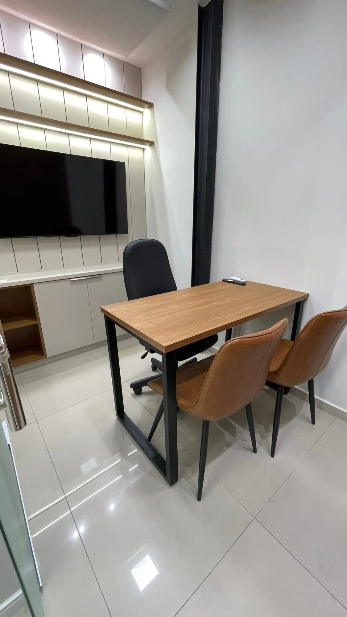 Sala privativa com mesa de madeira, cadeira executiva, duas cadeiras de couro caramelo e TV na parede ripada