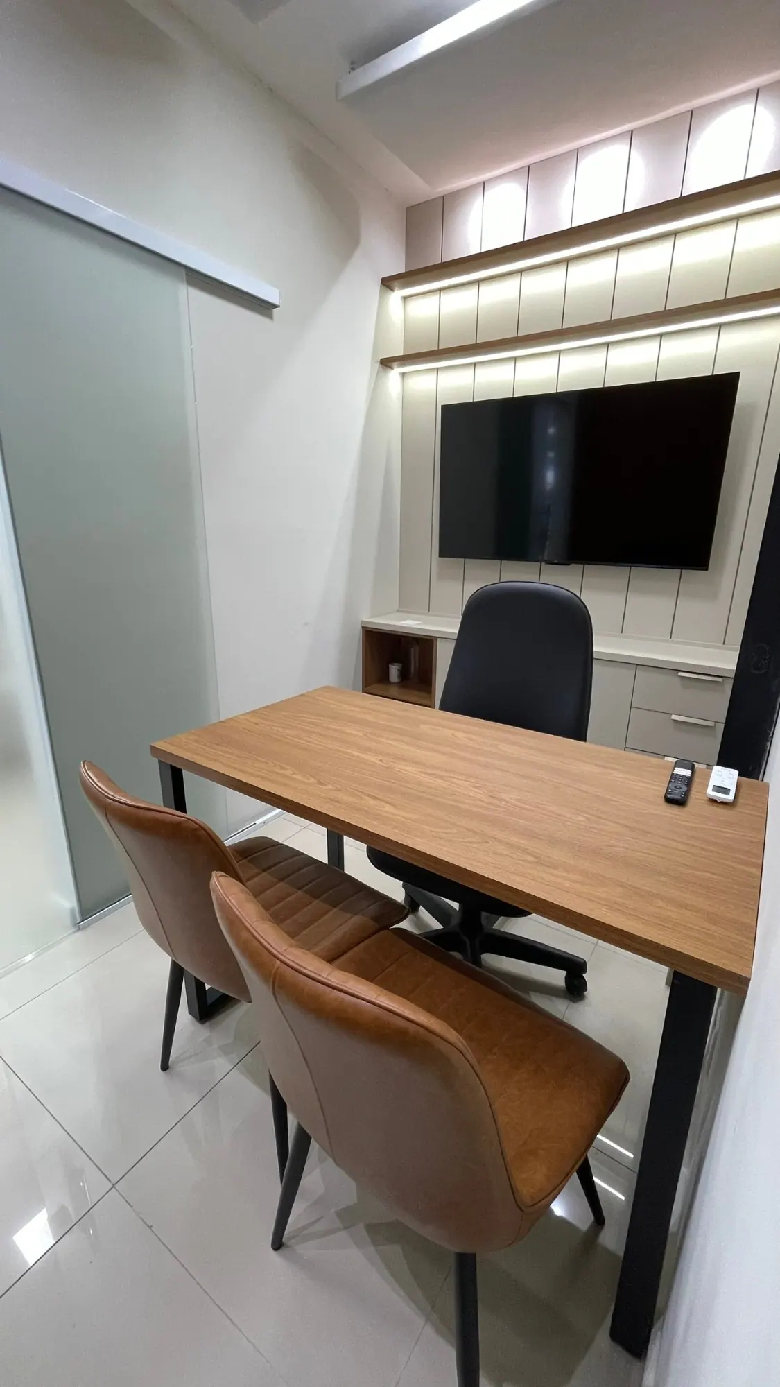 A mesma sala privativa vista da porta de vidro, com armário, prateleiras iluminadas e TV na parede