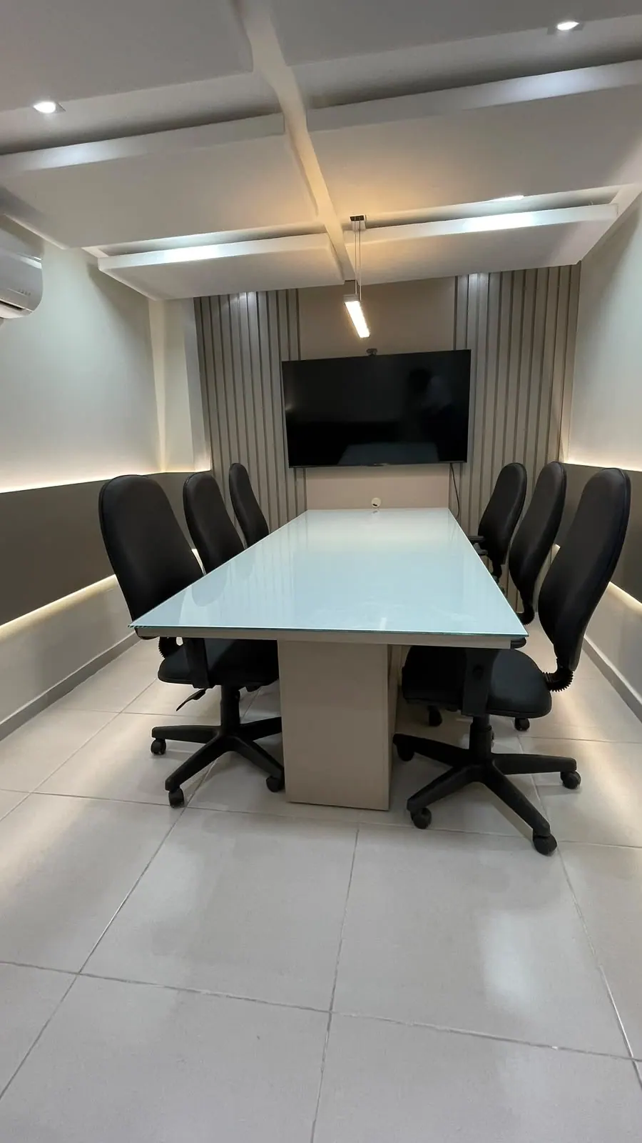 Sala de reunião com mesa de vidro, cadeiras pretas e TV na parede